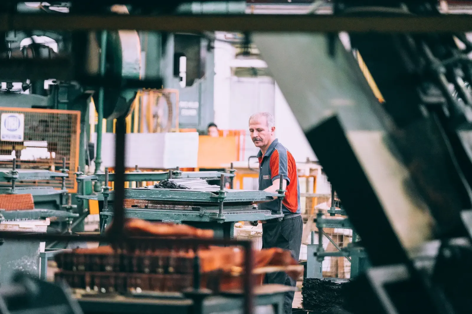 Man with a mustache in a red and gray shirt works amidst industrial machinery in a factory.
