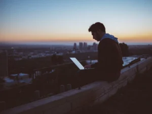 Silhouetted young man uses a laptop on a wall, overlooking a city skyline at sunset.