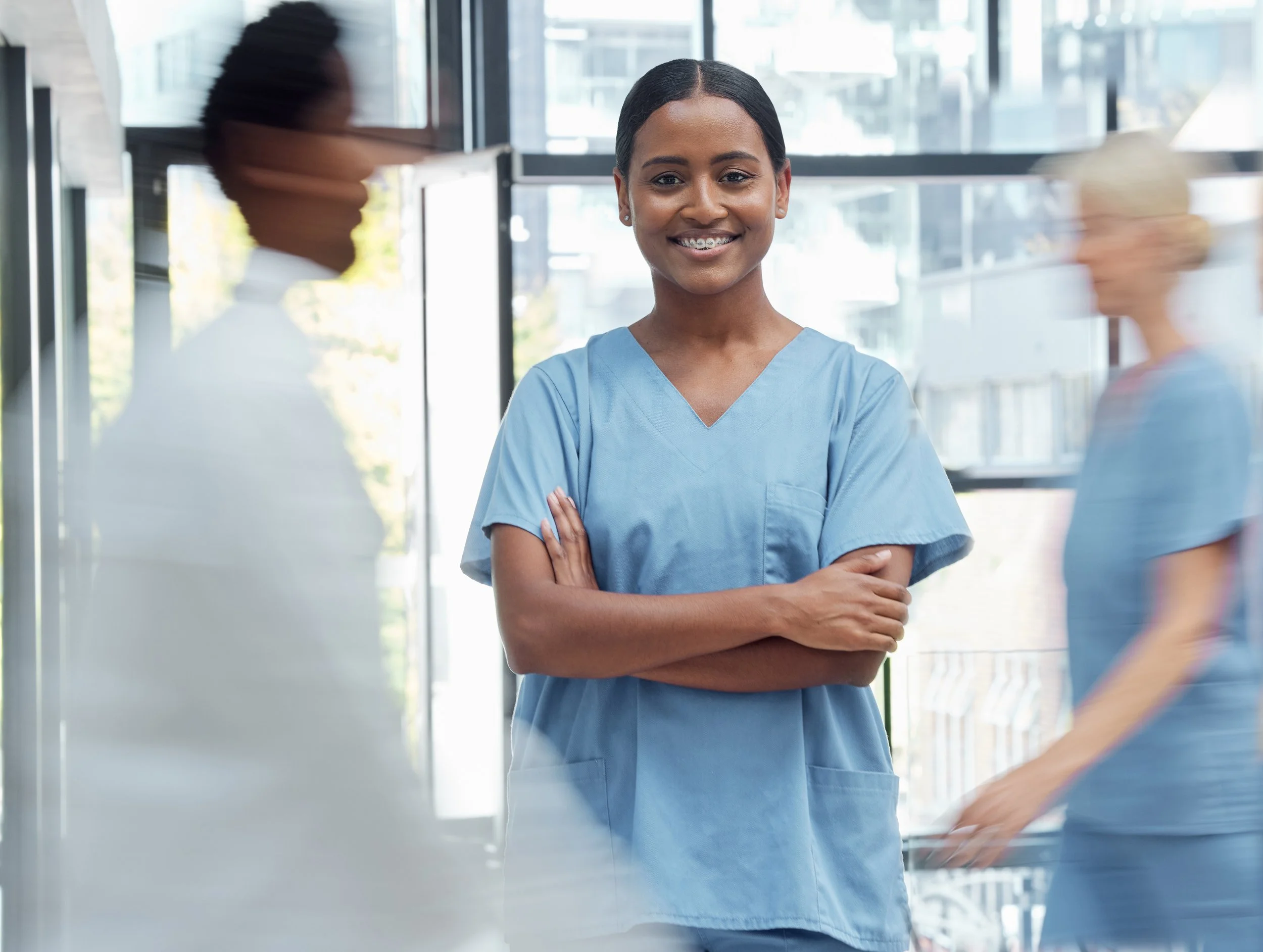 Smiling Black woman in blue scrubs with braces, arms crossed, with blurred medical staff.