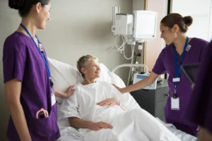 Two nurses in purple scrubs comfort a smiling elderly patient in a hospital bed.
